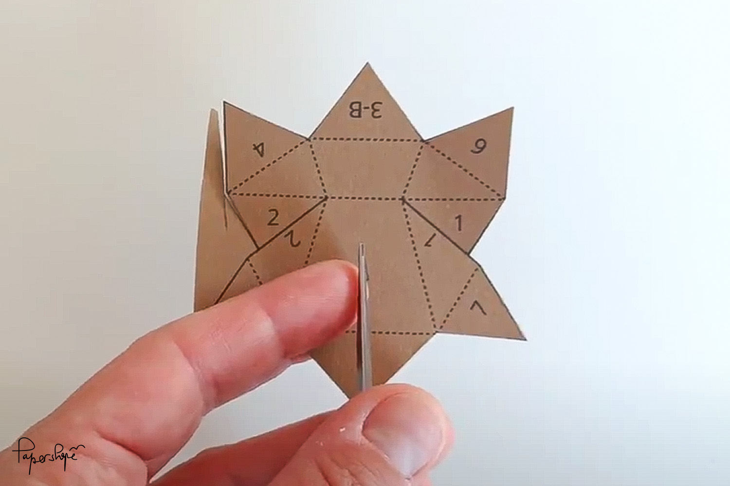 Hand schneidet mit Schere ausgeschnittenes Papercraft-Element aus braunem Papier mit markierten Laschen und gestrichelten Faltlinien