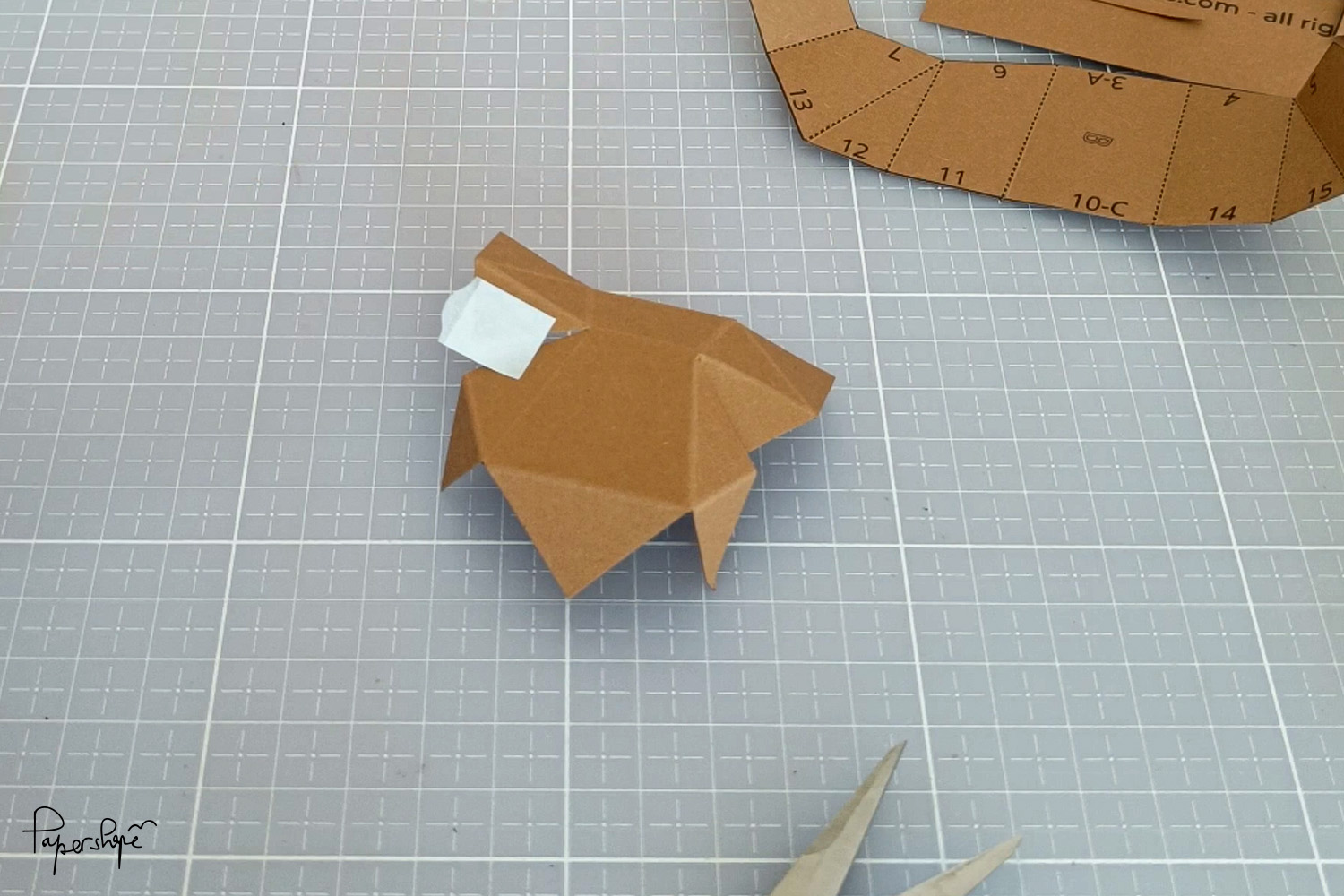 Teilweise zusammengeklebtes Papercraft-Element aus braunem Papier liegt auf Schneidematte; doppelseitiger Klebestreifen und weitere Bastelteile daneben