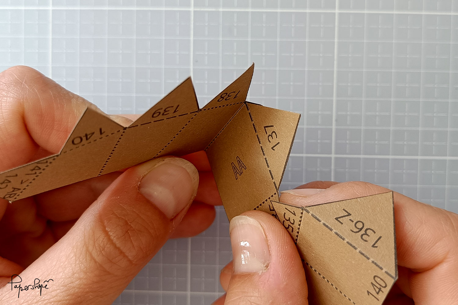 Zwei Hände falten eine nummerierte Papierform entlang einer gestrichelten Linie zu einer Bergfalte im Papercraft-Modell Dackel.