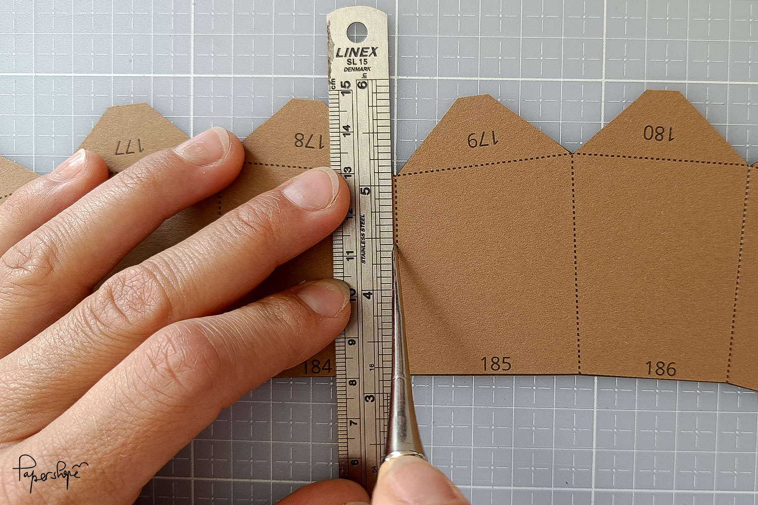 Hand benutzt ein Metalllineal und ein Falzwerkzeug, um entlang einer gestrichelten Linie auf braunem Bastelpapier eine präzise Falzlinie zu ziehen.