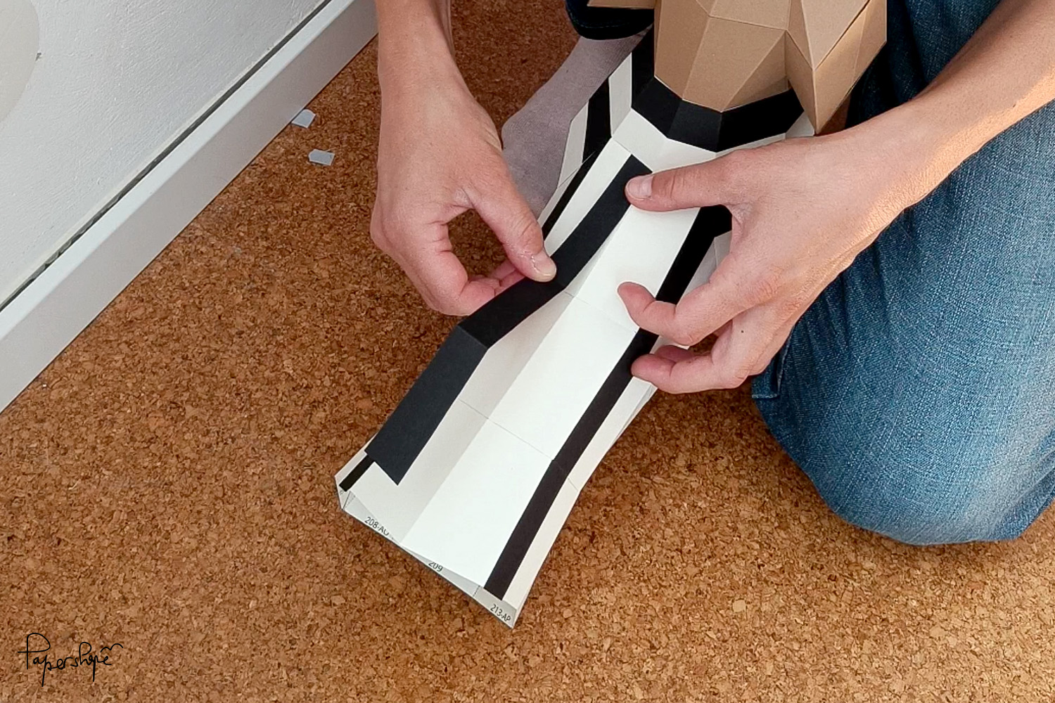 Zwei Hände kleben schwarze Papierstreifen auf die weiße Innenseite eines braunen, länglichen Papercraft-Dackelmodells