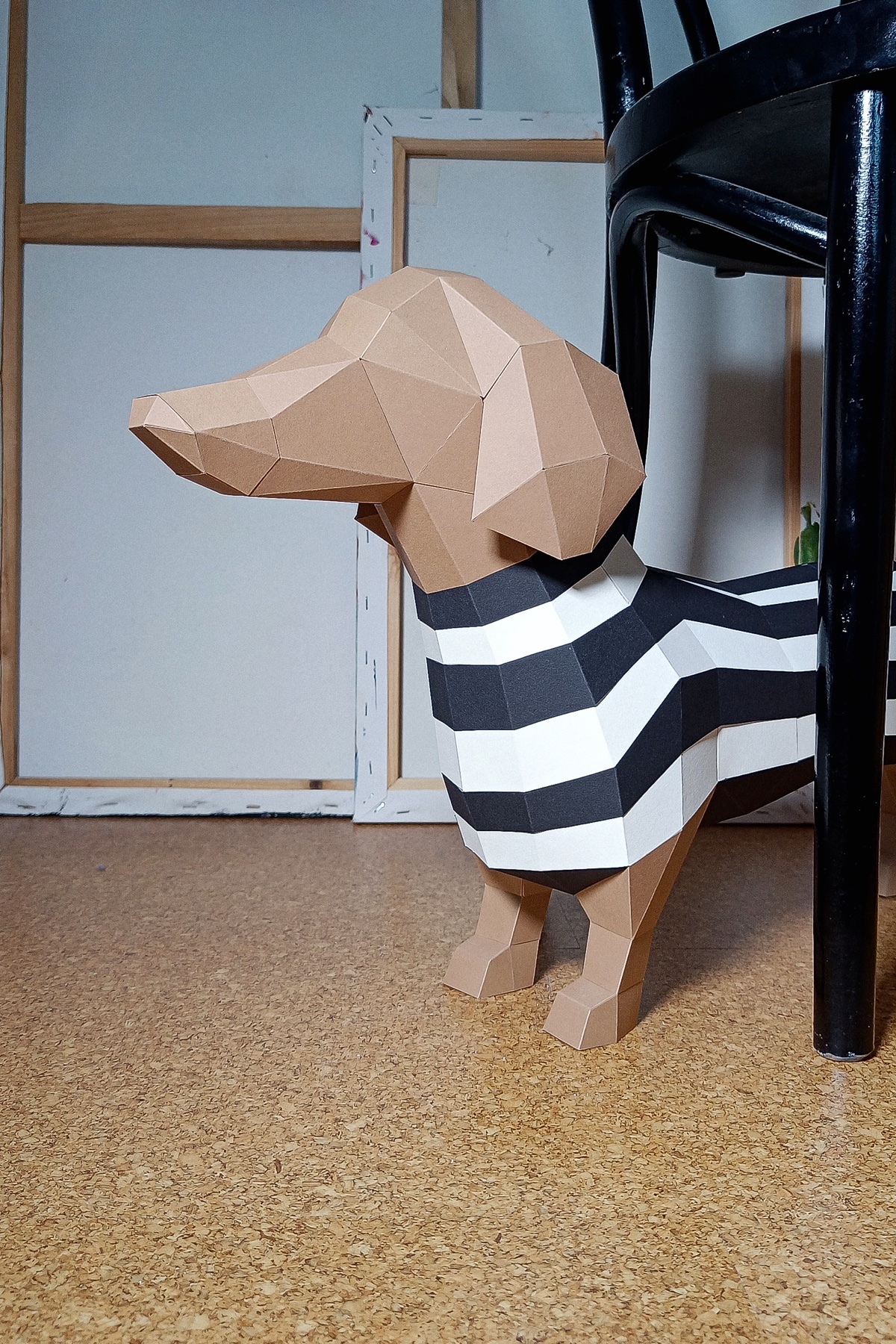 Papercraft Dackel aus braunem Tonkarton mit schwarz-weißem Streifenshirt, unter einem schwarzen Stuhl auf Korkboden vor Leinwandrahmen im Atelier.