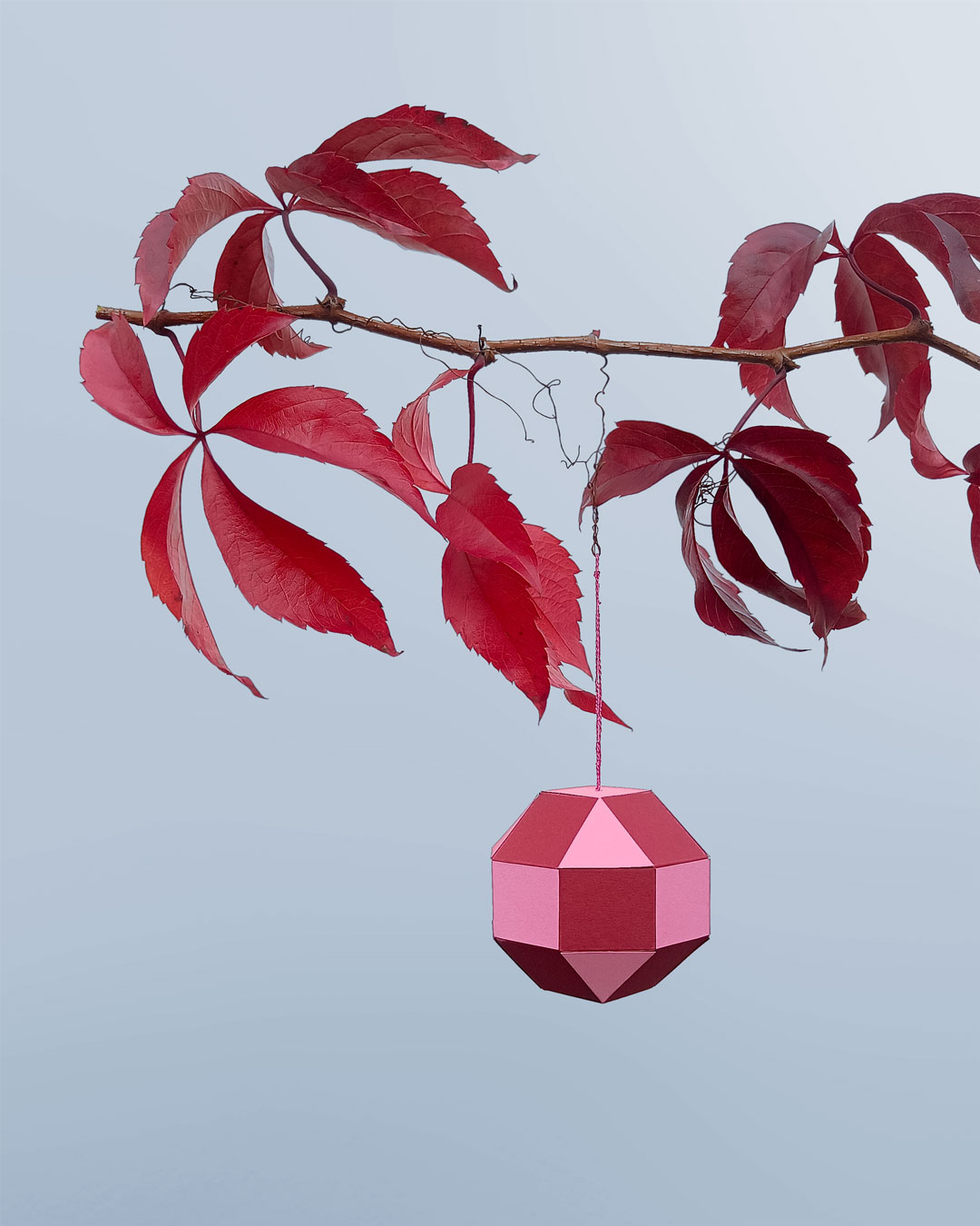 Red geometric paper pendant hanging from a branch with red leaves - minimalist paper art.