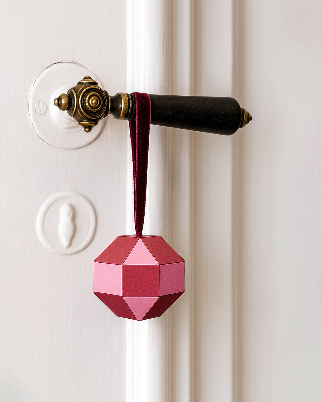 Geometric paper pendant in cherry red and eosin with velvet ribbon hanging from an antique door handle - elegant Christmas decor.