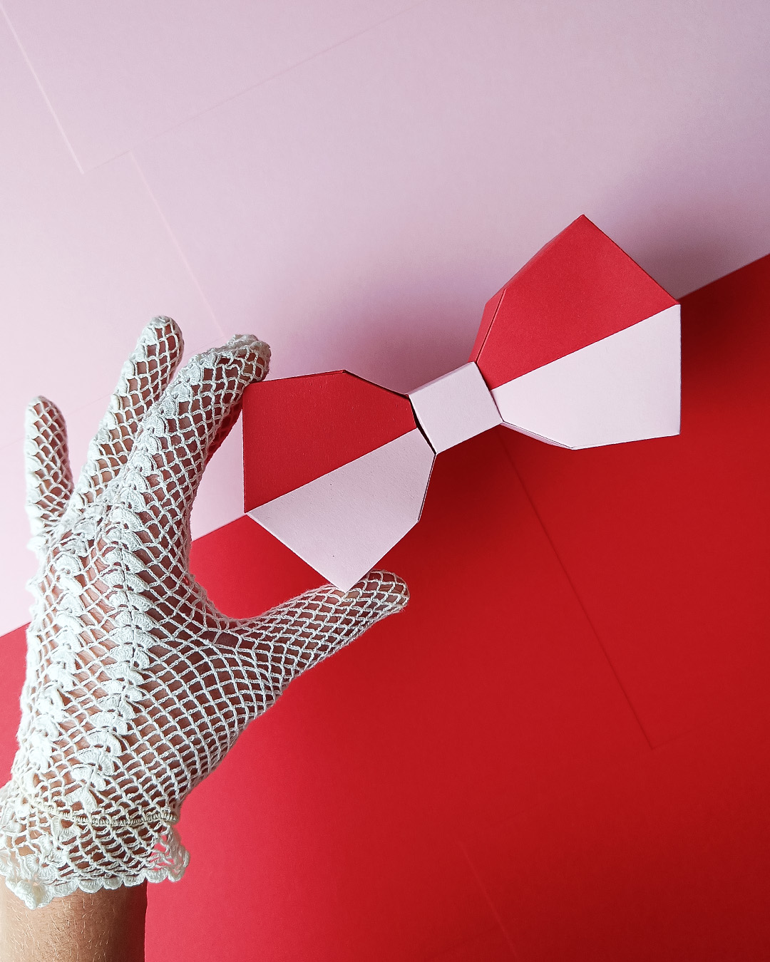 Hand mit Handschuh hält eine 3D Low-Poly Papierschleife - vom flachen Bastelbogen zum fertigen Objekt, roter Hintergrund.
