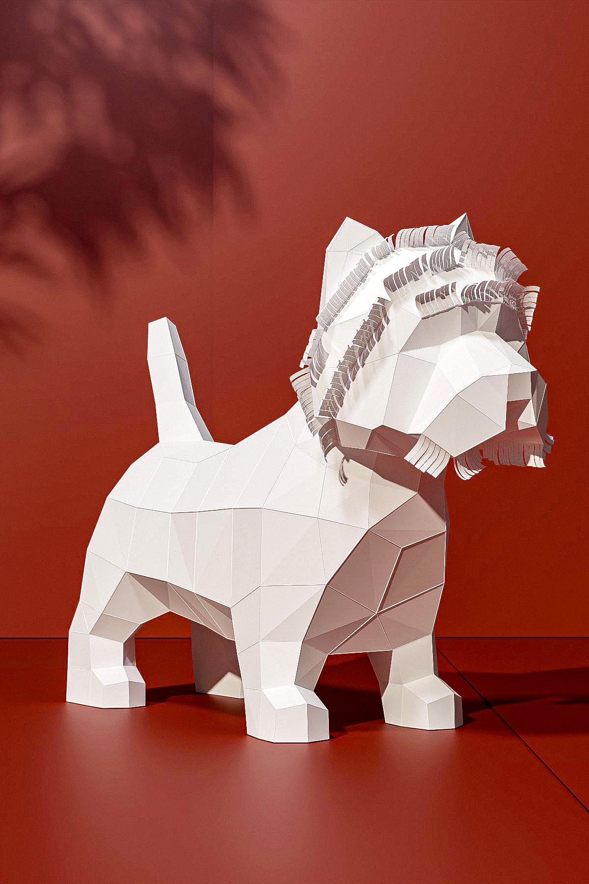 Papercraft Westie Elisabetta made of white cardstock in low poly style, in side profile with its tail up and cut fringes on its head, against a terracotta red wall with plant shadows.
